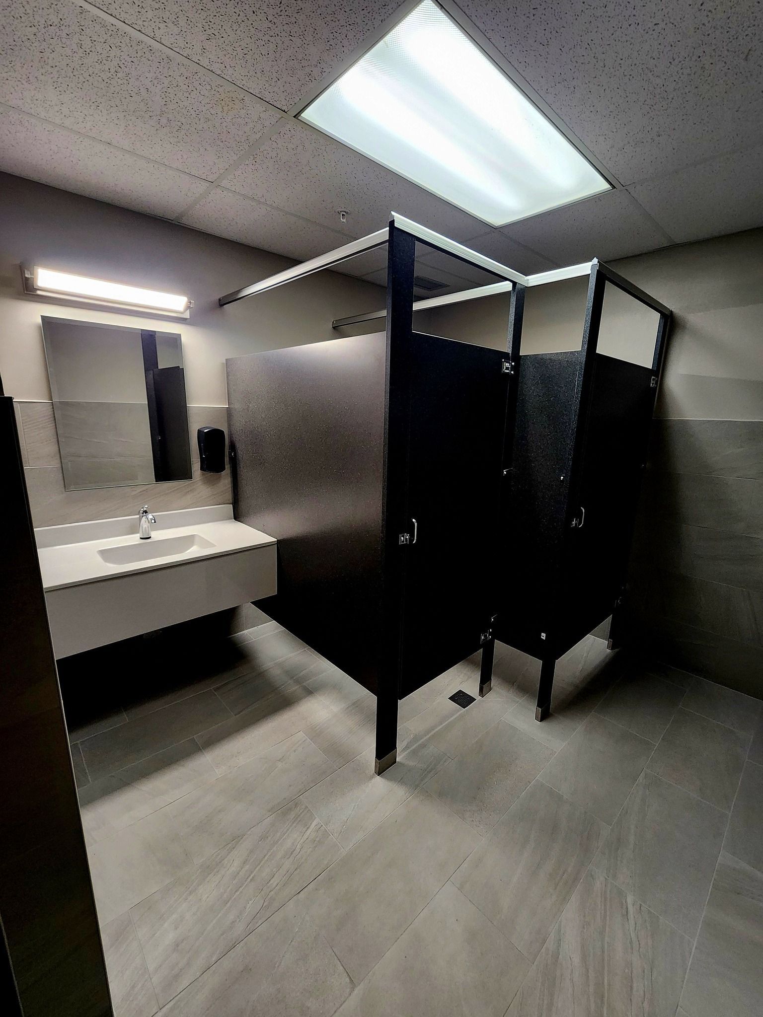 Public restroom with black stall partitions, white sink counter, and fluorescent ceiling lights.