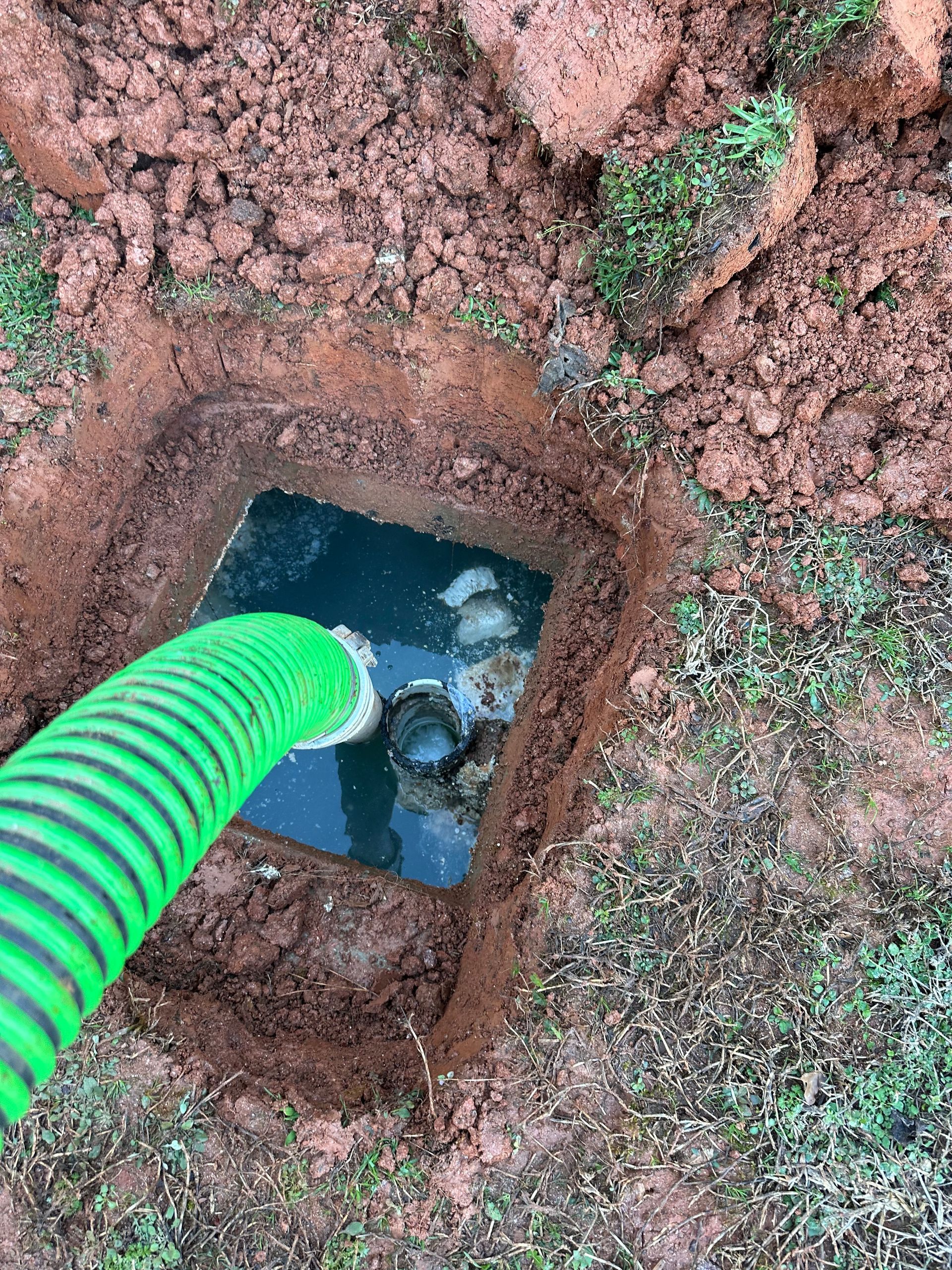 Green hose pumping liquid from a square hole dug in red soil, surrounded by grass.