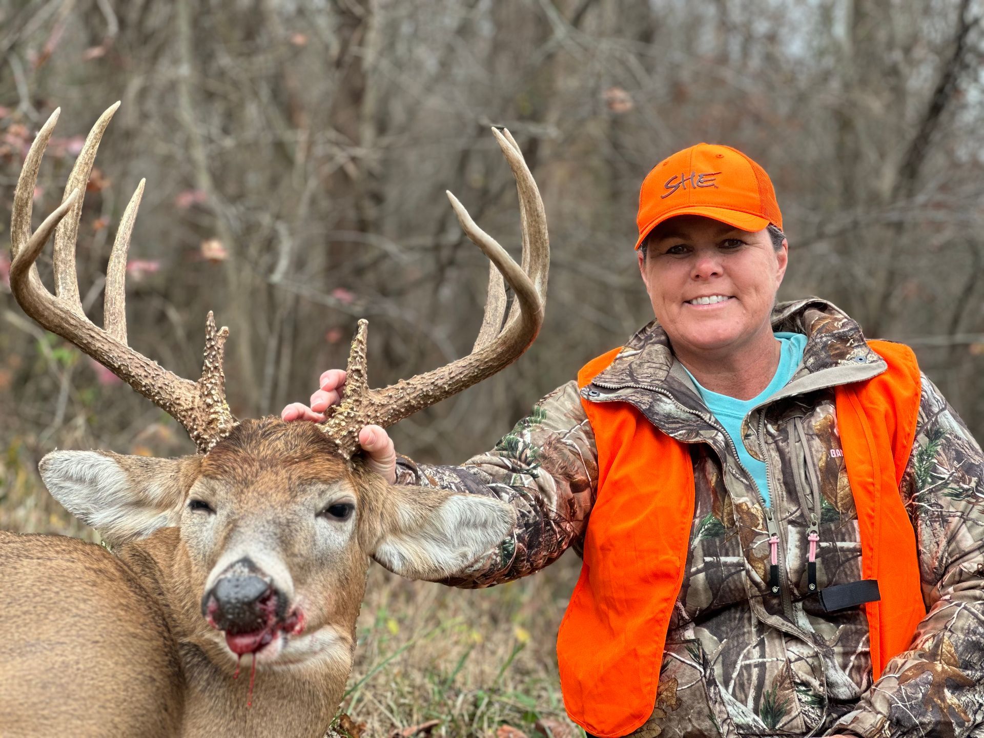 Woman in hunting gear smiles, posing with large buck in wooded area. She wears orange vest and hat, touches deer's antlers.