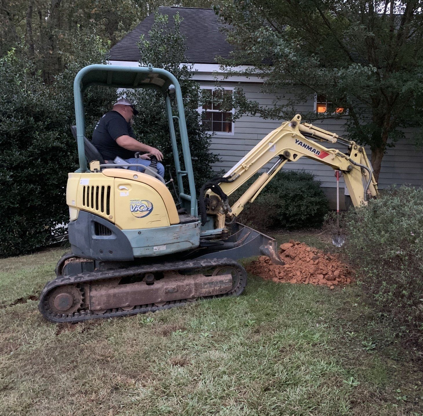 Person operating a yellow mini excavator in a grassy yard near a house, digging into a dirt pile.