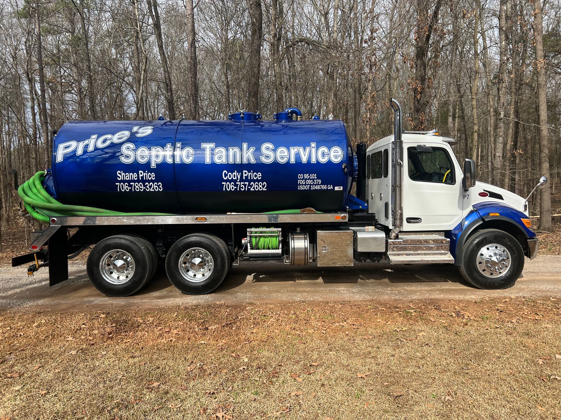 Blue and white septic tank truck with green hose in wooded area, branded 