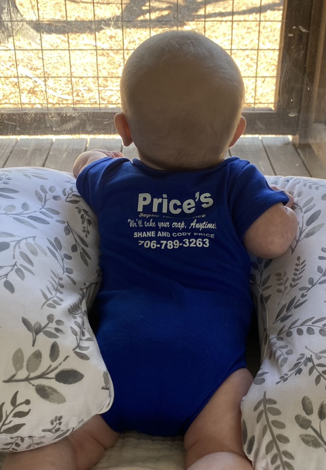 Baby in a blue onesie, looking out a window while propped on a patterned pillow.