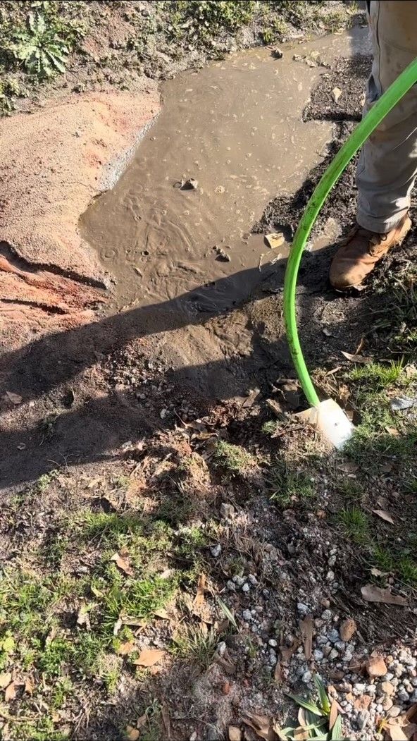Person watering muddy puddle with green hose outdoors.