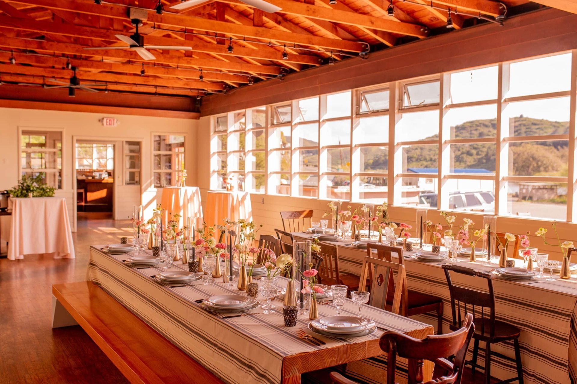 Wedding reception with long tables, floral centerpieces, large windows overlooking a waterfront. Warm, golden light.