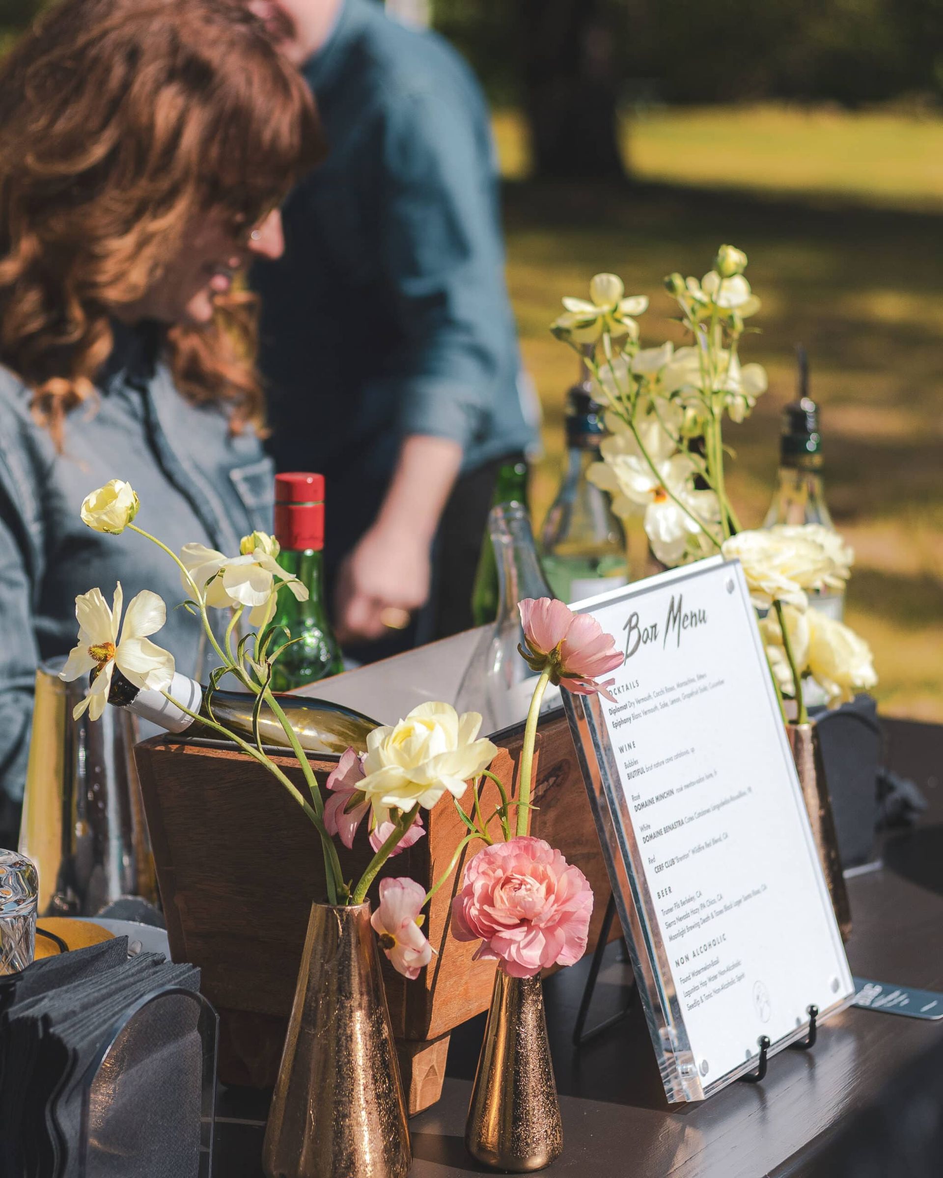 Cocktail bar setup with flowers, menu, bottles. Person in denim shirt behind bar. Outdoor setting.