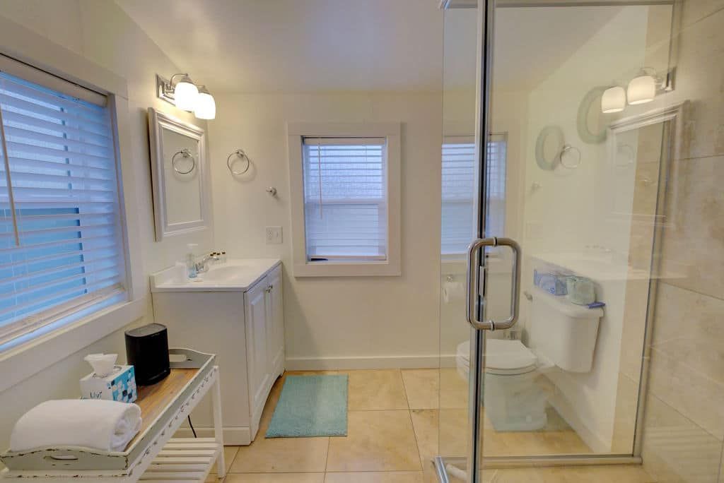 Bathroom with white vanity, shower, and toilet. Light-colored tile floor, windows with blinds, and white walls.