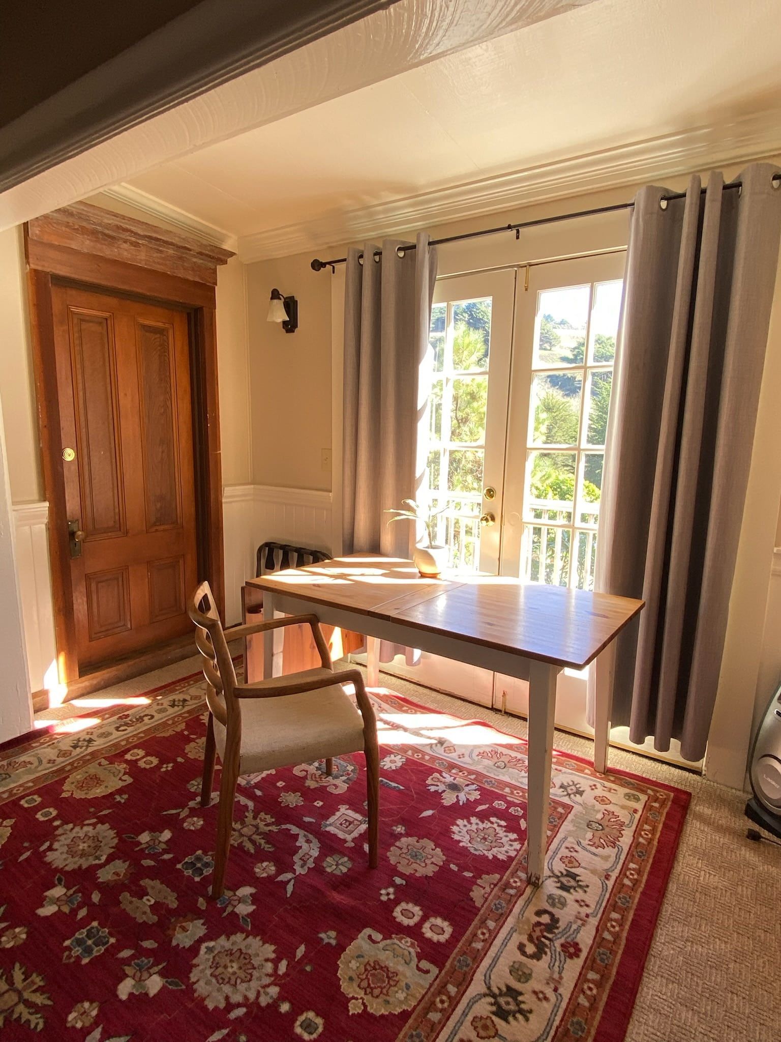 Room with a table and chair on a red floral rug, near a door and window with light coming through.