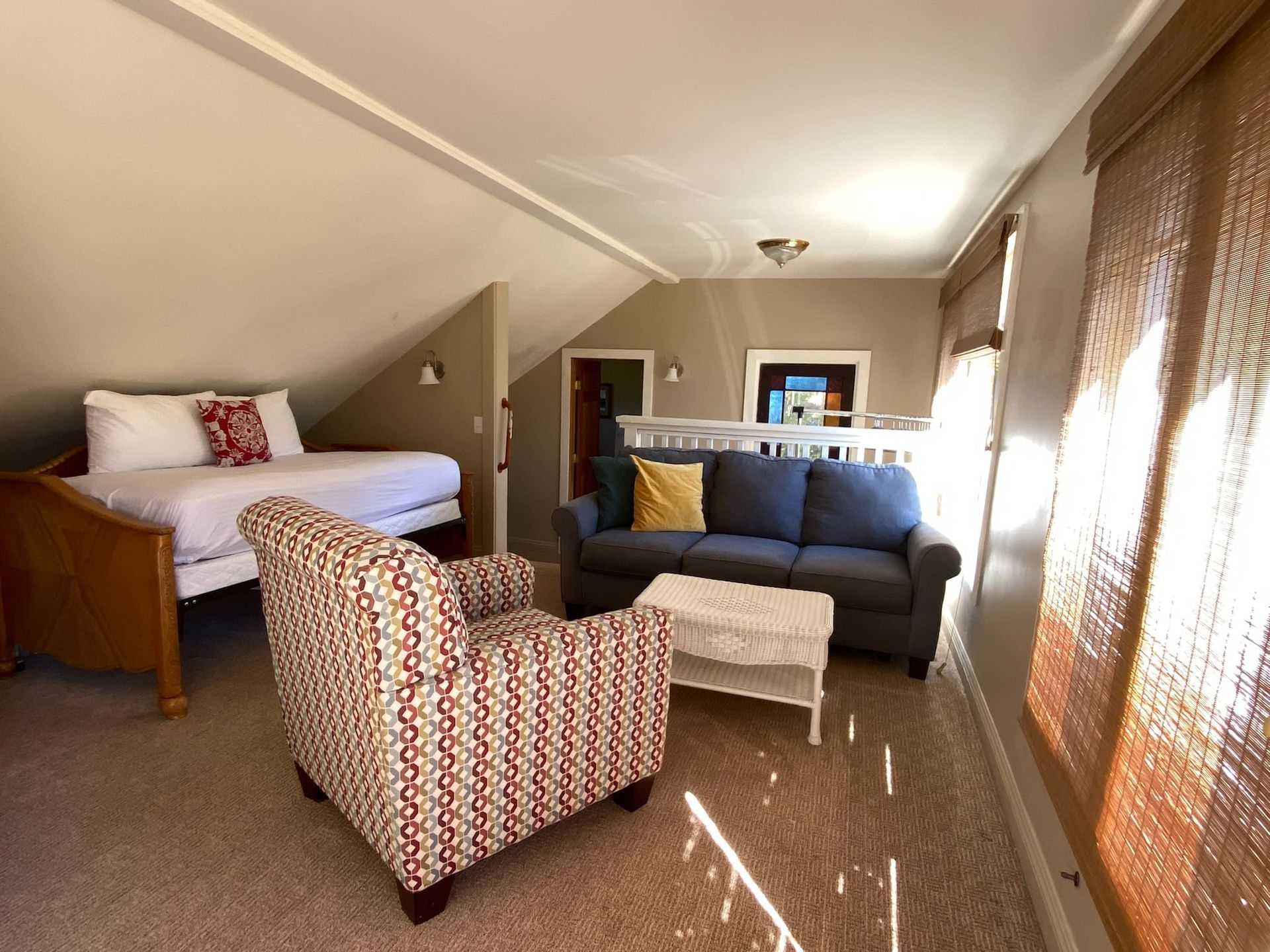 Loft room with bed, sofa, patterned armchair, and woven window shades; natural light.