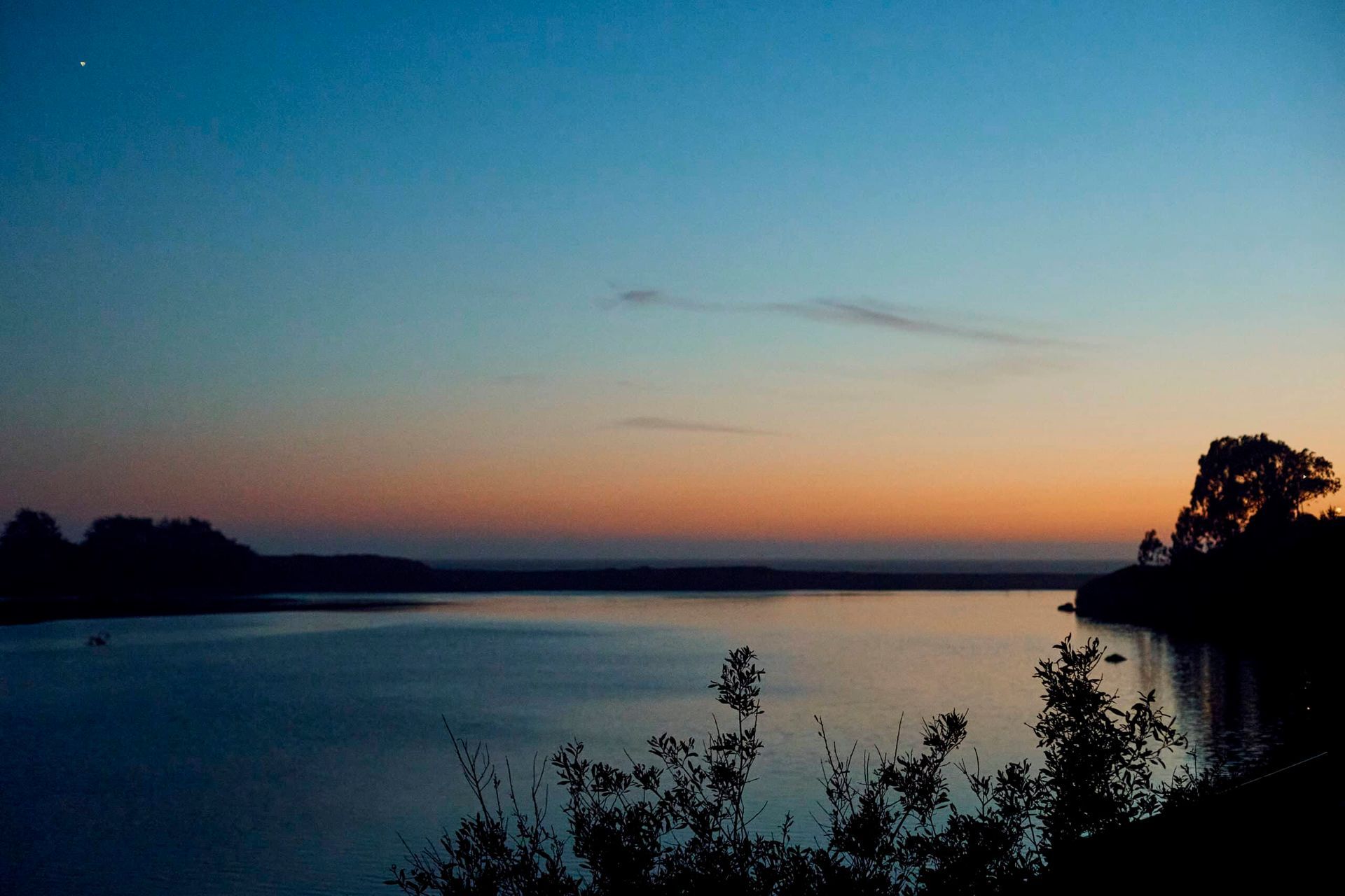Sunset over a calm lake, with trees silhouetted against a gradient sky of blue, orange, and purple.