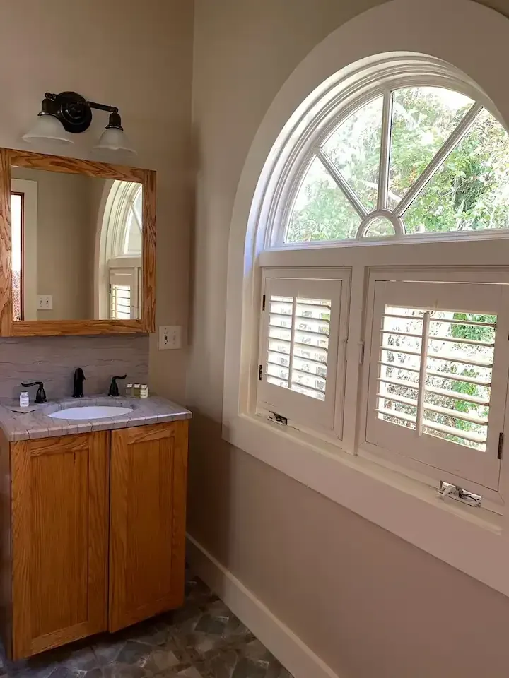 Bathroom with wooden vanity, arched window with shutters, and a mirror.