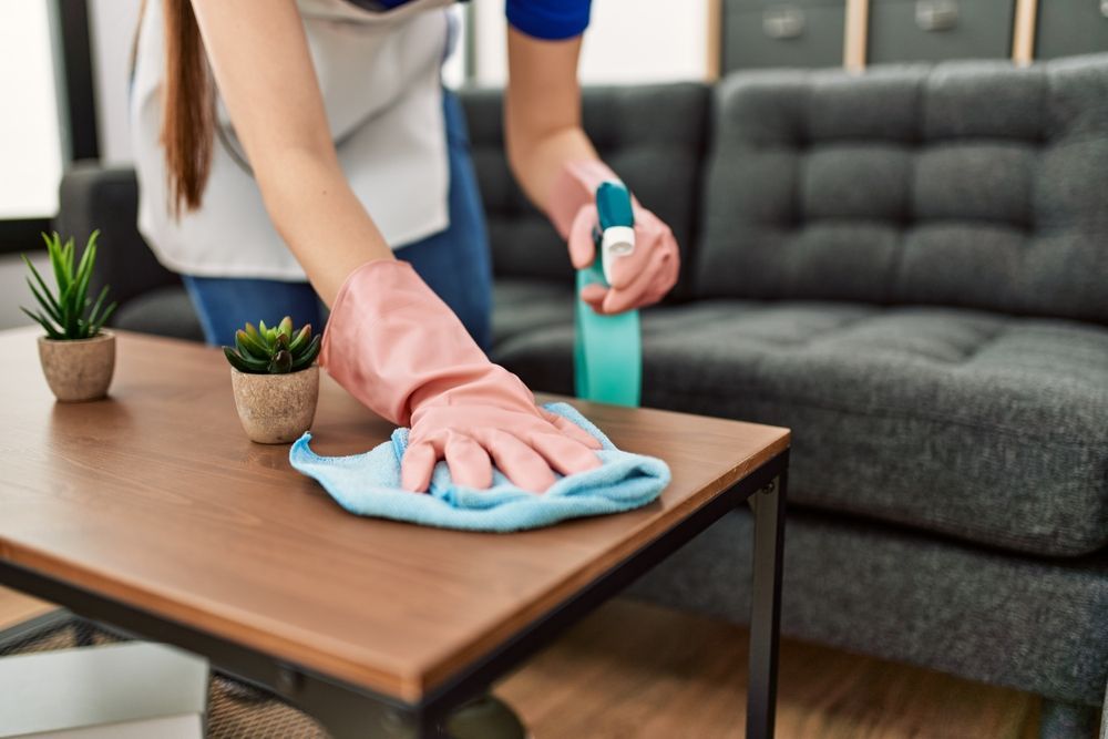 Person cleaning a wooden table with a spray bottle and cloth, next to a couch.