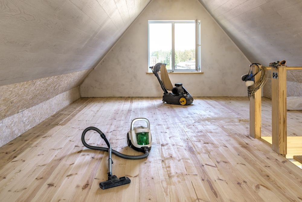 Attic with light wood floor, vacuum, and floor sander near a window. Wooden railing on right.