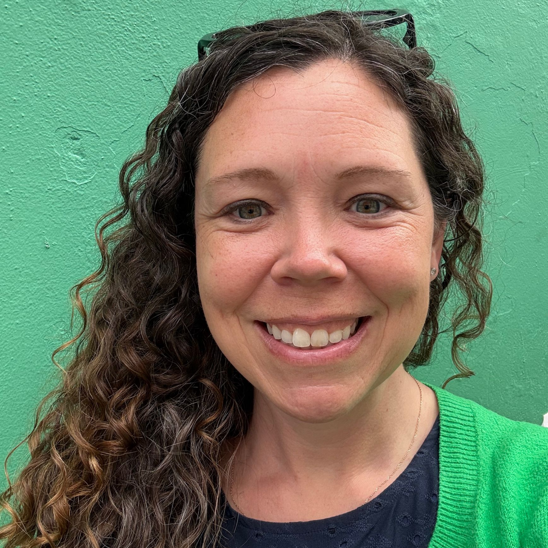 A person with long, curly brown hair and a friendly smile, wearing a green cardigan against a matching green background.