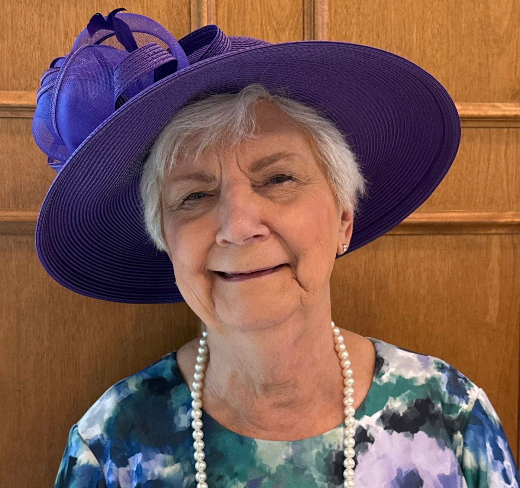 A person wearing a wide-brimmed purple hat and a pearl necklace, smiling against a wood-paneled wall.