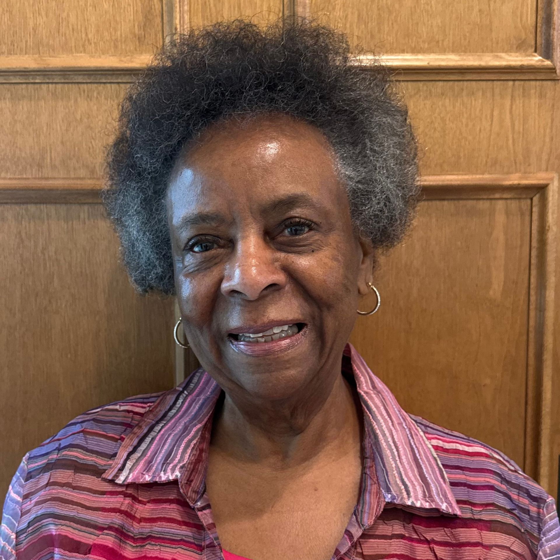 A person wearing a striped, multi-colored button-up shirt and hoop earrings, smiling in front of a wooden paneled wall.