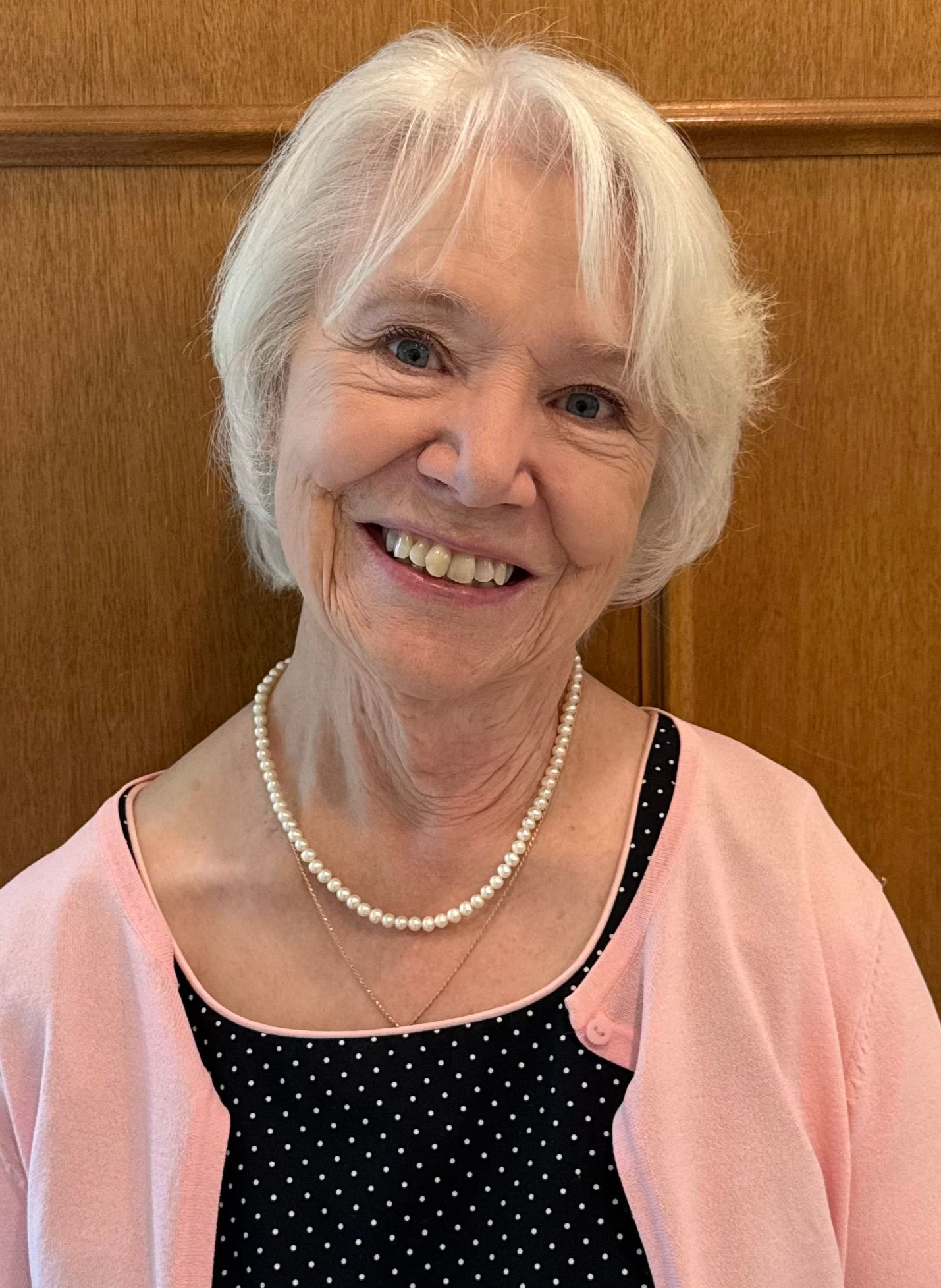 Smiling person with white hair, wearing a pearl necklace, a black polka-dot top, and a light pink cardigan.