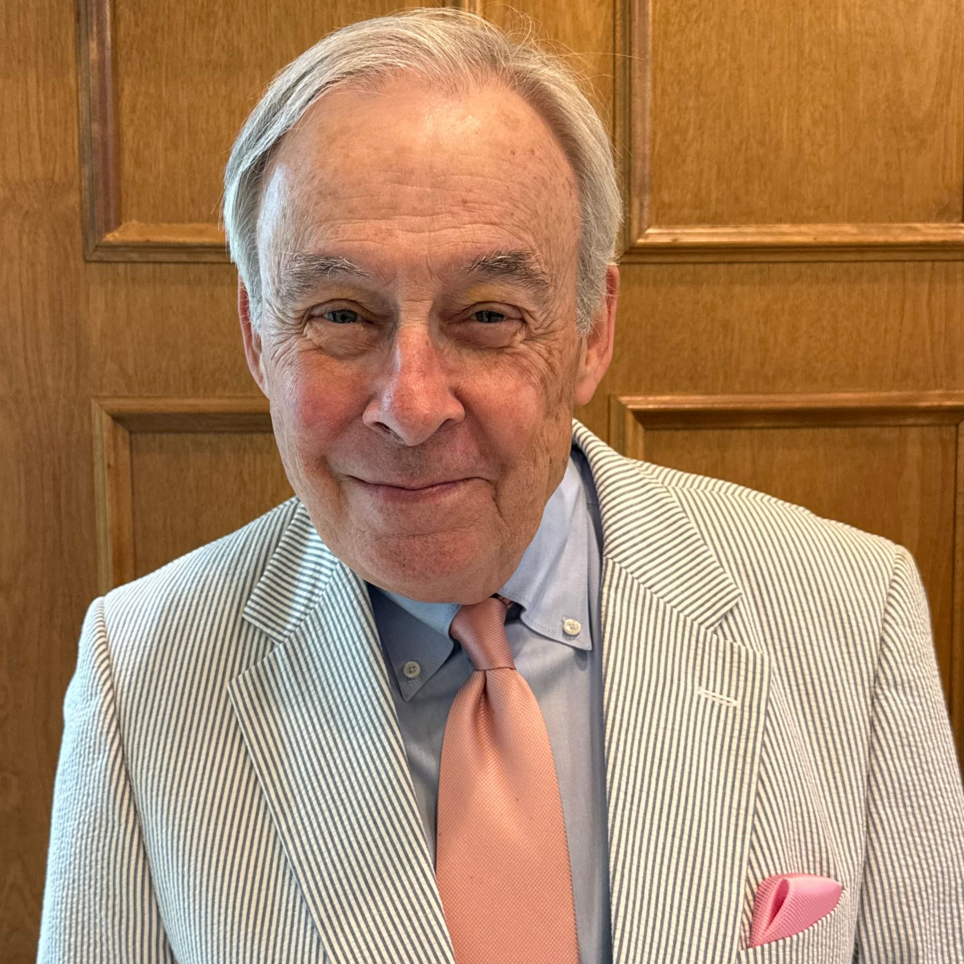 A person wearing a striped seersucker blazer, blue shirt, and pink tie stands in front of a wood-paneled wall.