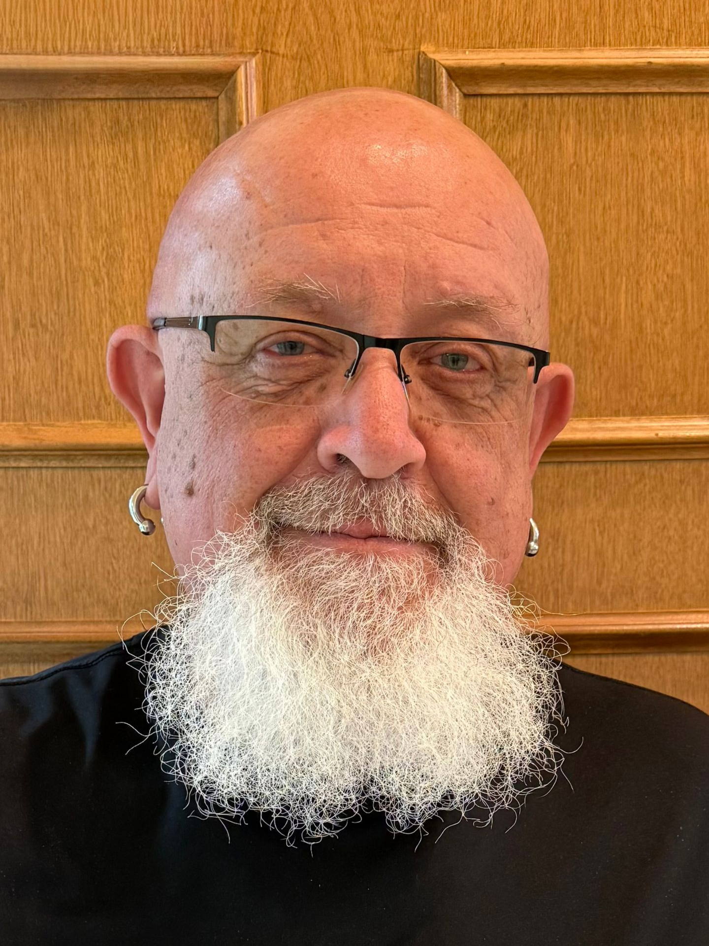 A person with a white beard and glasses wearing hoop earrings, posed against a paneled wood wall.