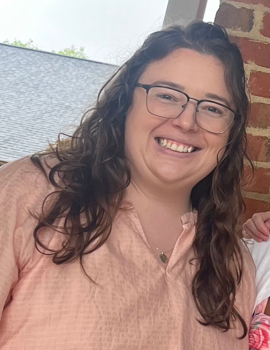 A person with shoulder-length brown hair and glasses smiles in front of a red brick wall.