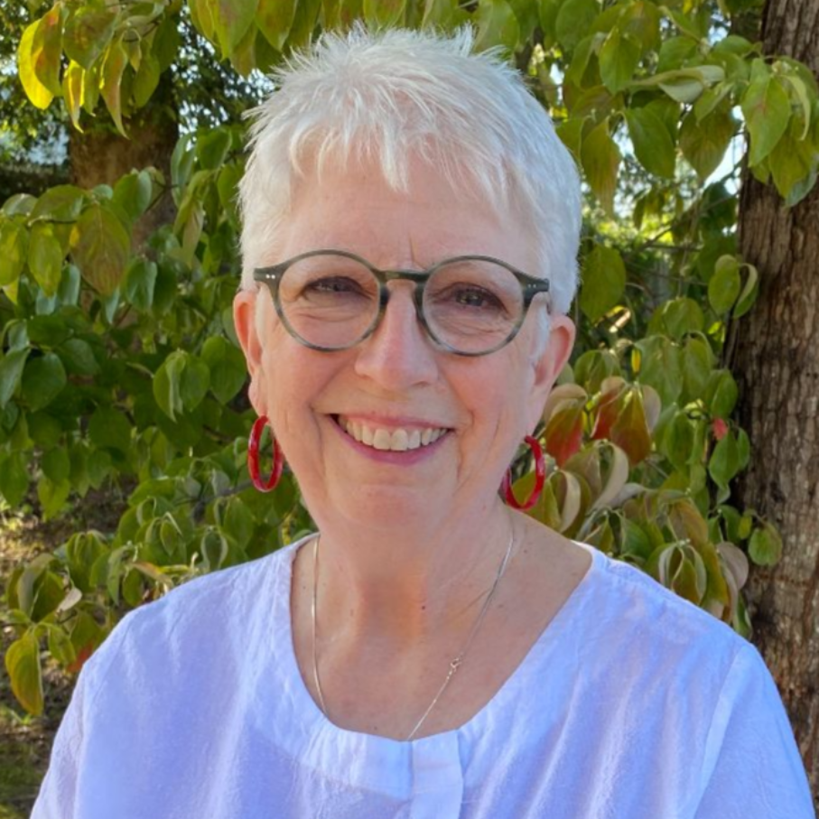 A person with short white hair and round glasses smiles while wearing a white shirt and red hoop earrings in a garden.