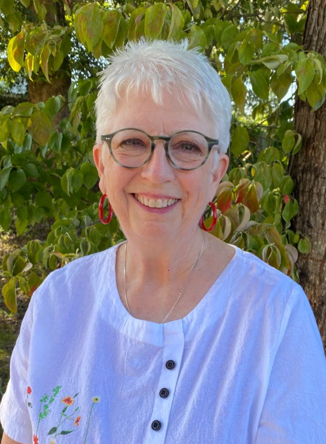 A smiling person with short white hair and round glasses, wearing a white shirt, outdoors in front of a leafy tree.