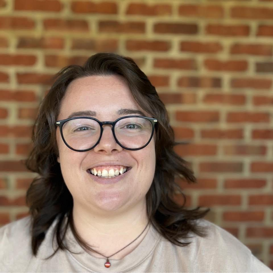 A person with shoulder-length brown hair and glasses smiles in front of a red brick wall.