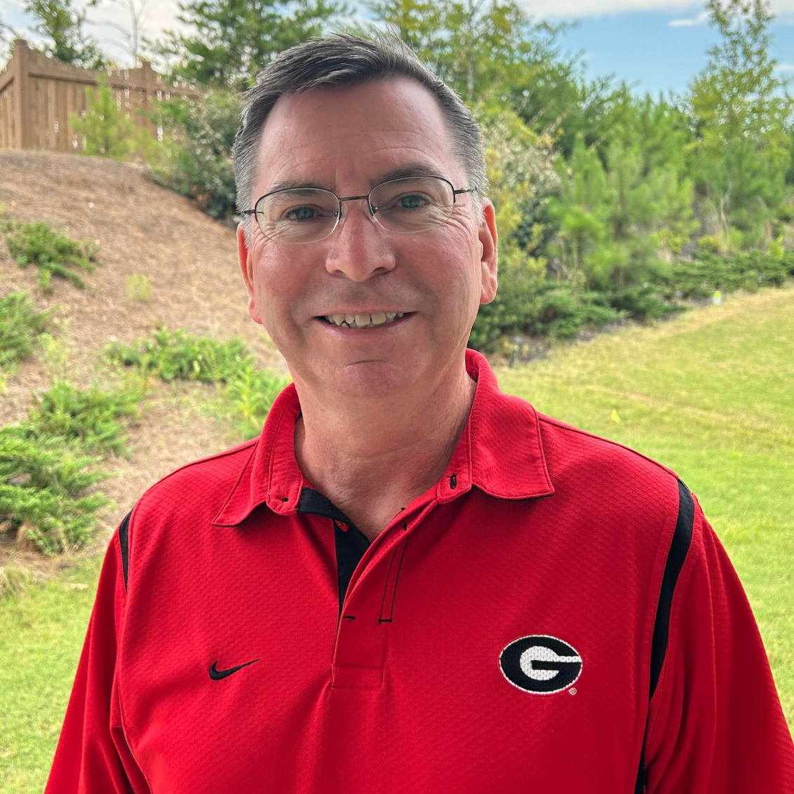 A smiling person wearing a red University of Georgia collared shirt with a Nike logo, standing outdoors near greenery.
