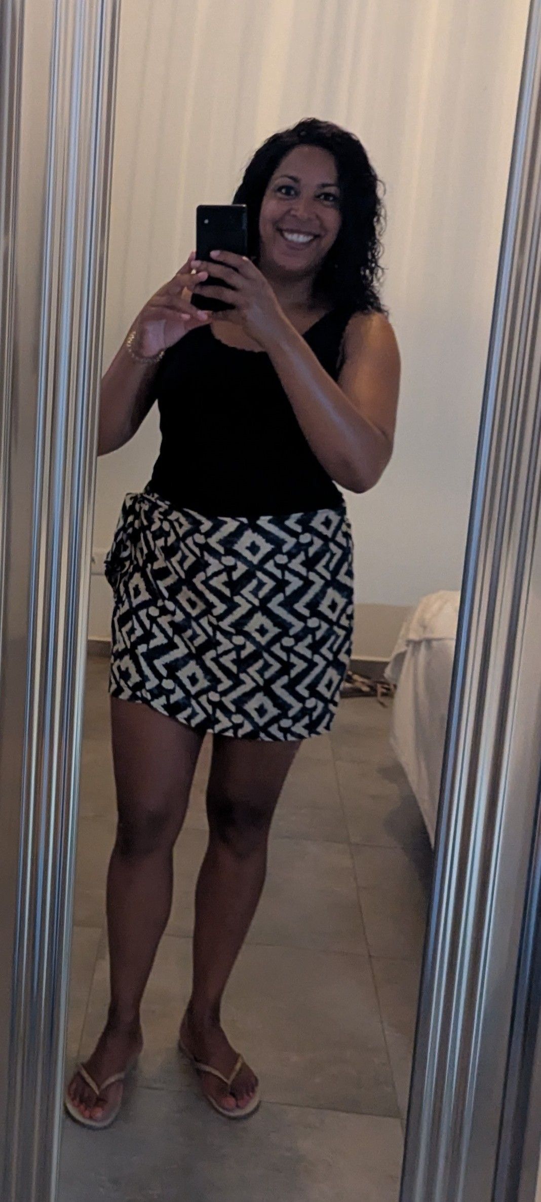 Spiegelselfie van een lachend persoon in een zwarte top en een rok met patroon, staand in een betegelde kamer.