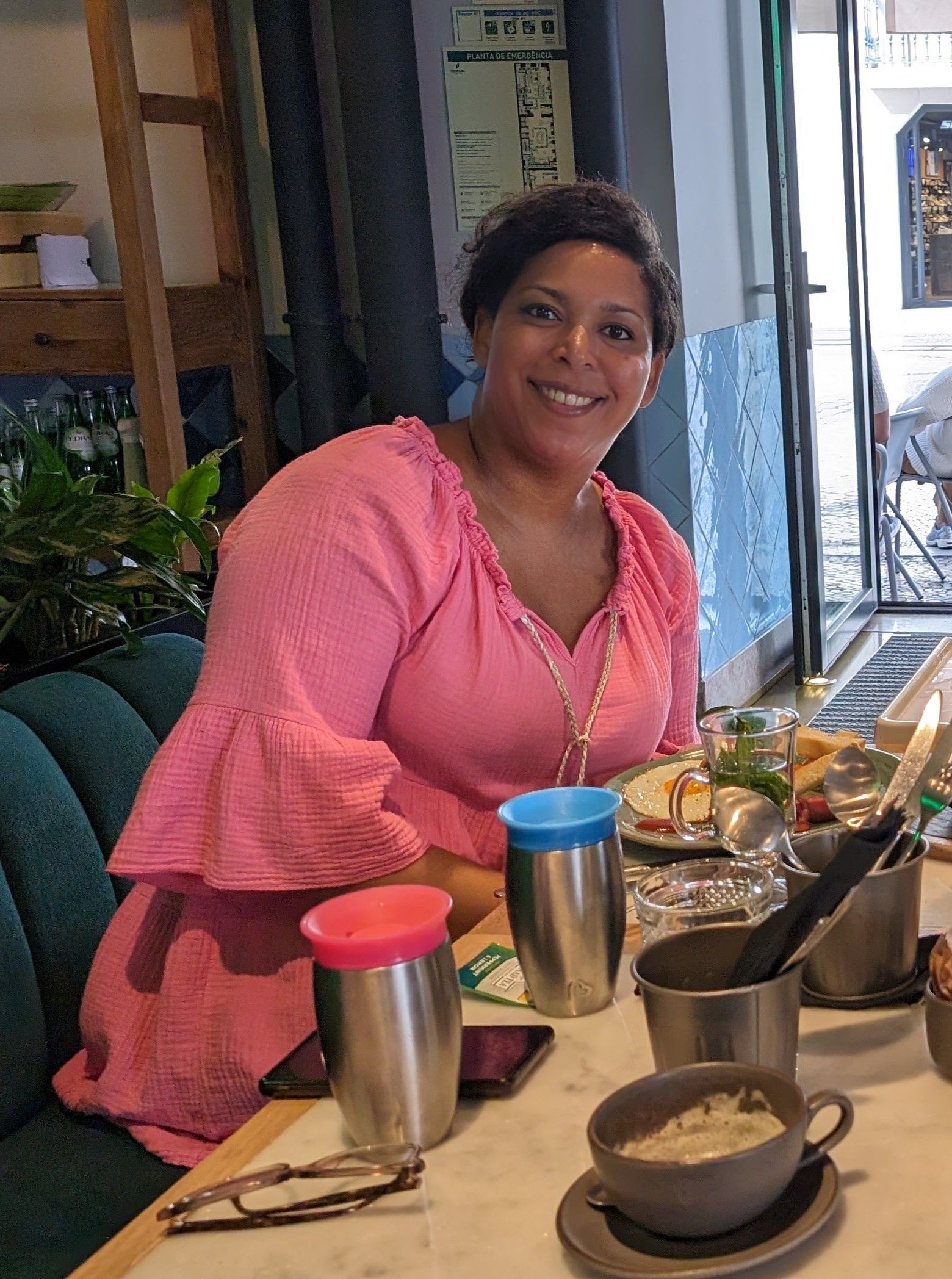 Een lachend persoon in een roze topje zit aan een restauranttafel met eten en drinken.