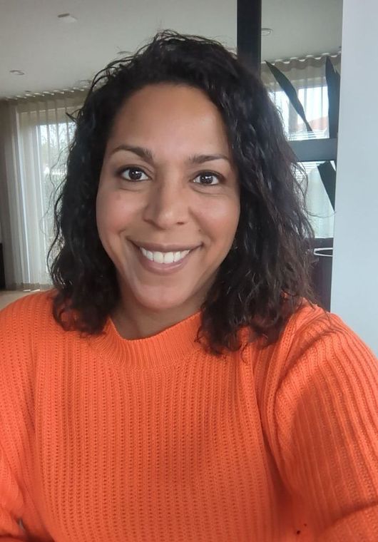 Een lachend persoon in een oranje trui maakt een selfie binnenshuis bij een raam.