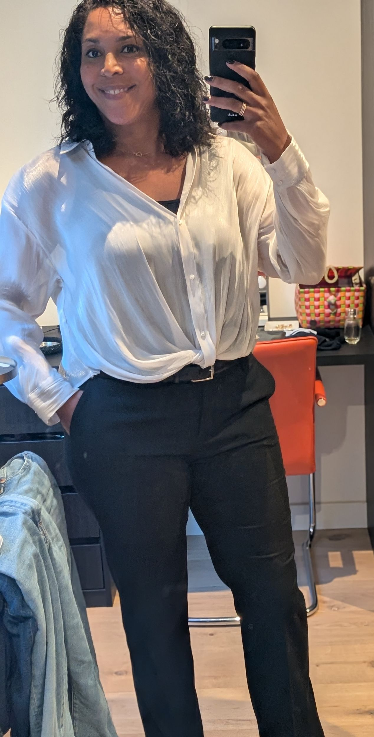 Vrouw in een witte blouse en donkere broek maakt een selfie in de spiegel, staand in een lichte kamer.