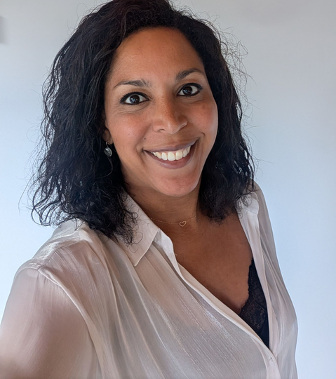 Een lachend persoon met krullend zwart haar in een lichte blouse tegen een effen muur.