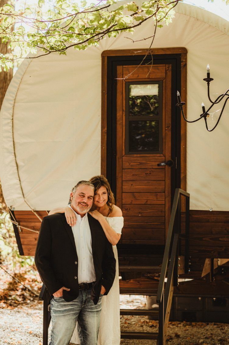 Couple standing beside a wooden door on a white trailer under tree branches