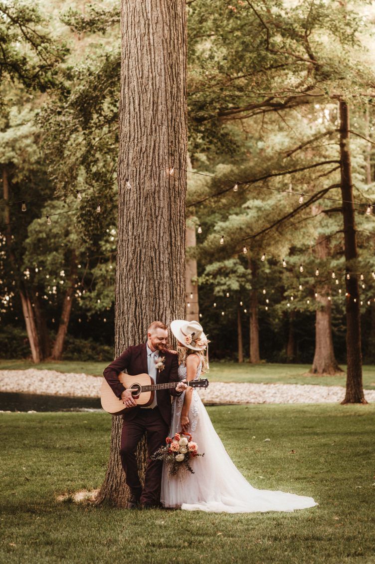 Bride and groom embracing by a large tree in a wooded park, holding flowers and a guitar