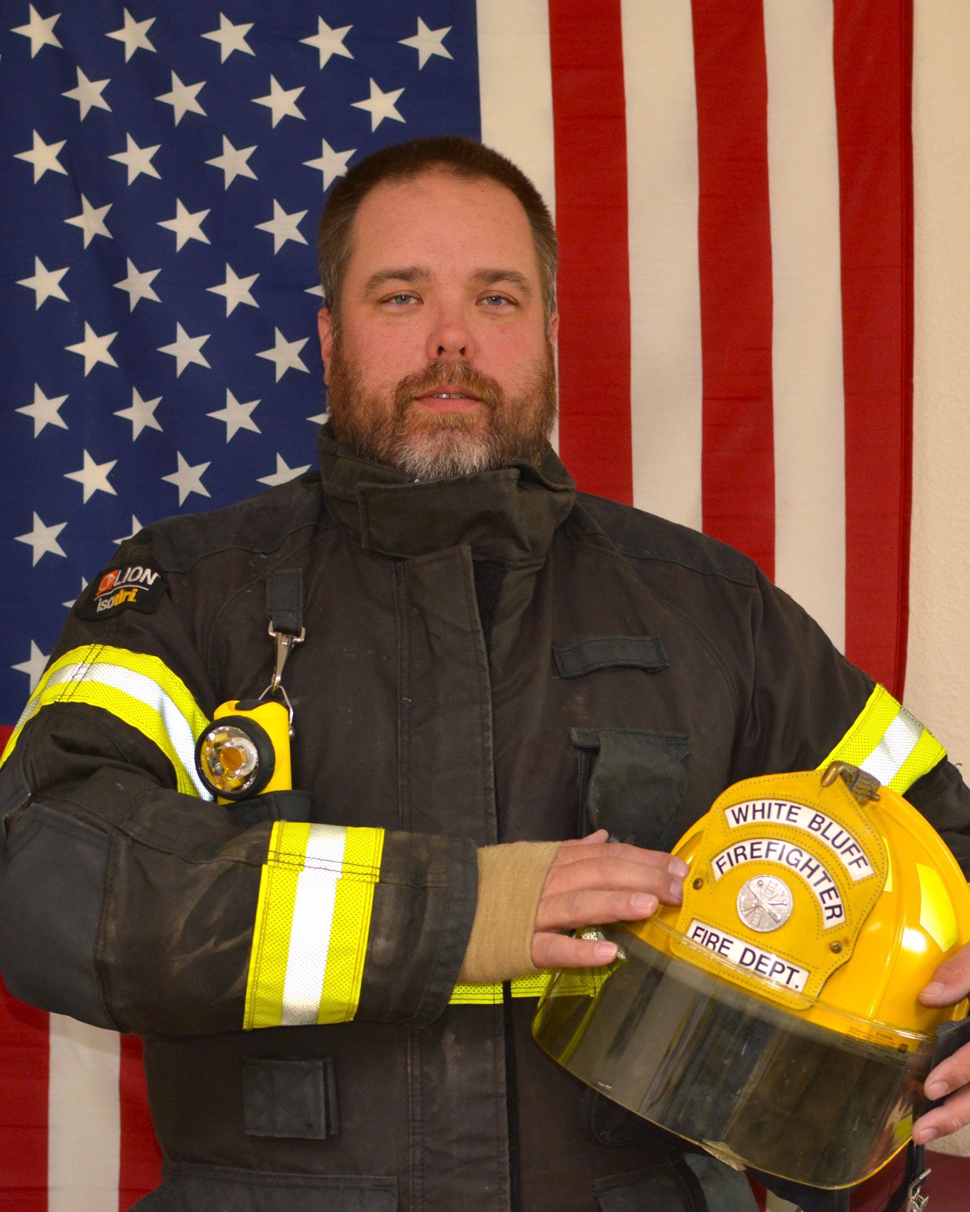 A man in a fireman 's uniform is holding a helmet and a flashlight in front of an american flag.