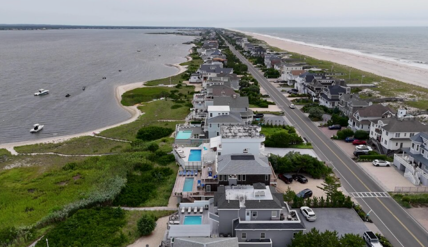 Aerial view of a coastal neighborhood beside a beach and bay, with houses, road, and greenery along the shoreline