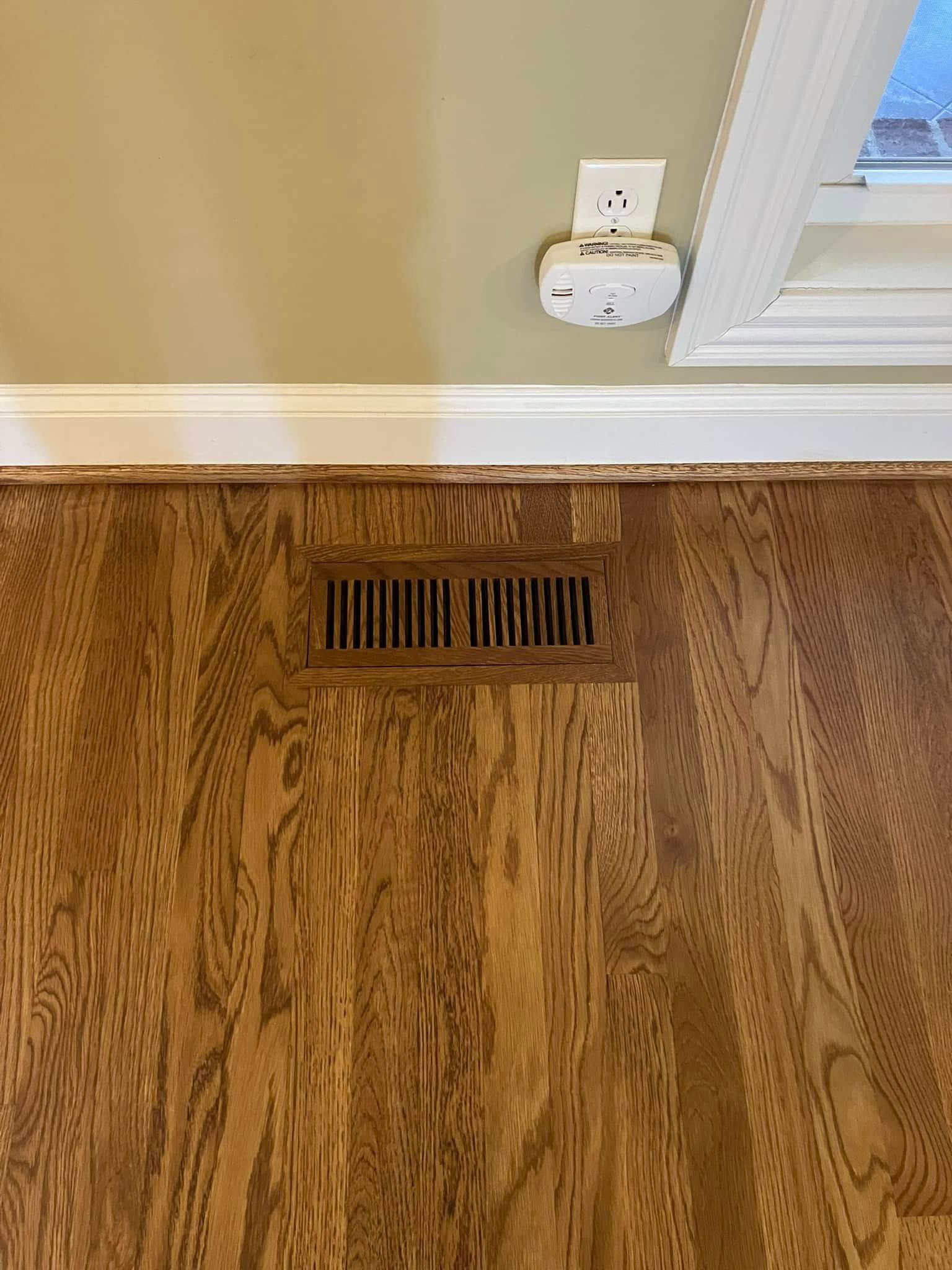 Floor vent in hardwood floor below a wall outlet and window trim
