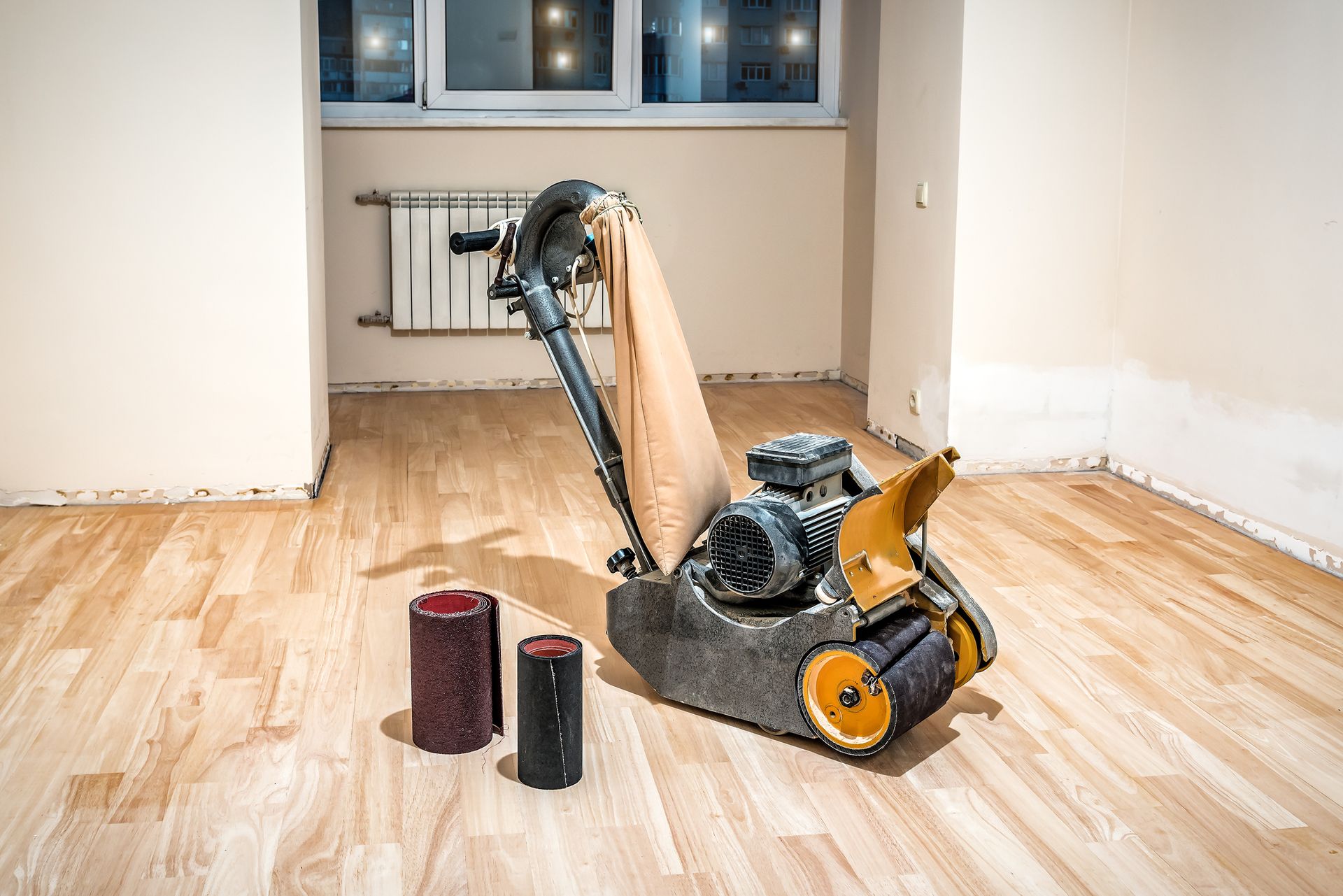 Floor sander sanding a wooden room floor with dust bags nearby, windows and radiator in the background
