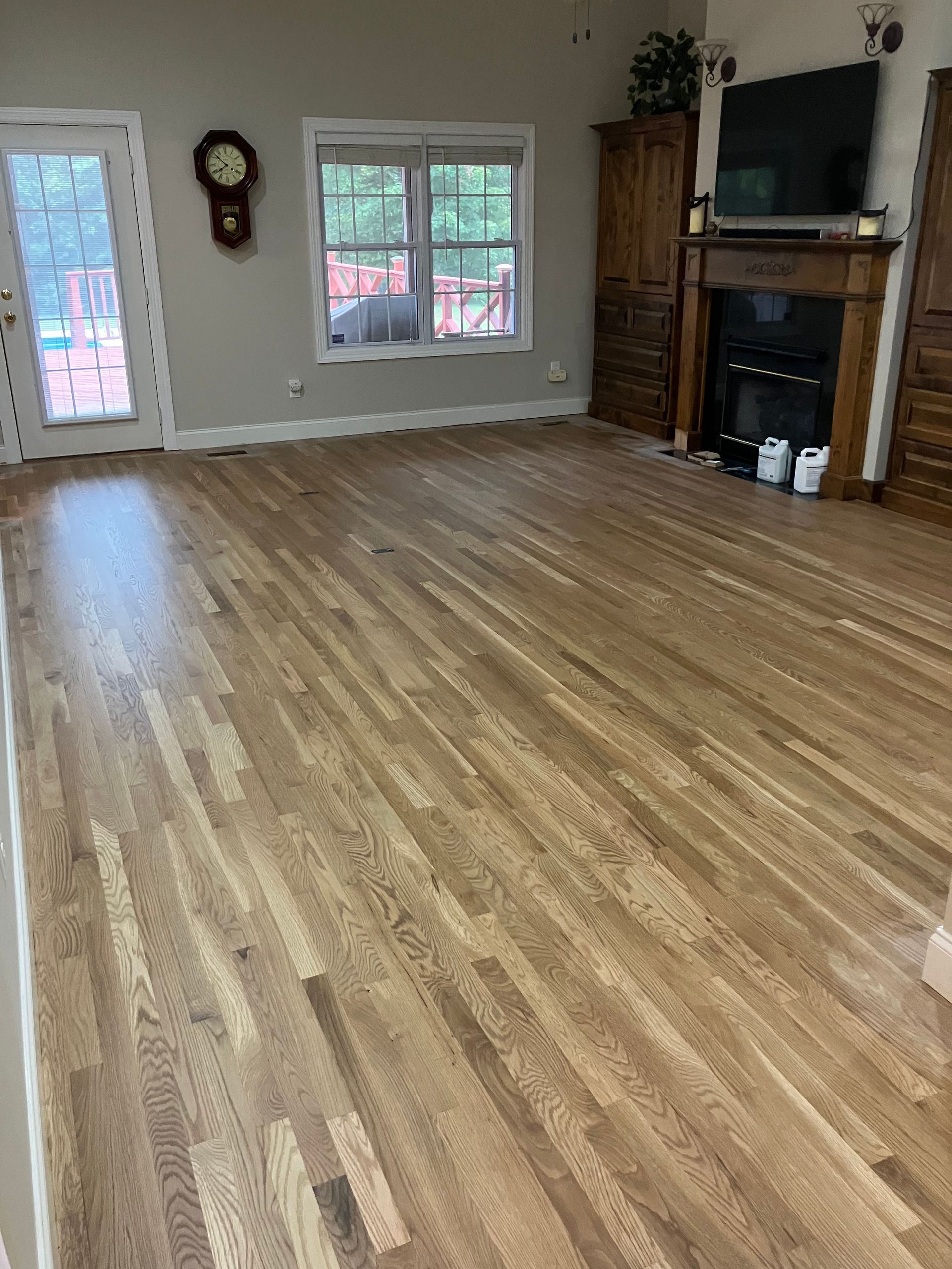 Empty living room with wood flooring, fireplace, wall clock, windows, and a TV on the right.