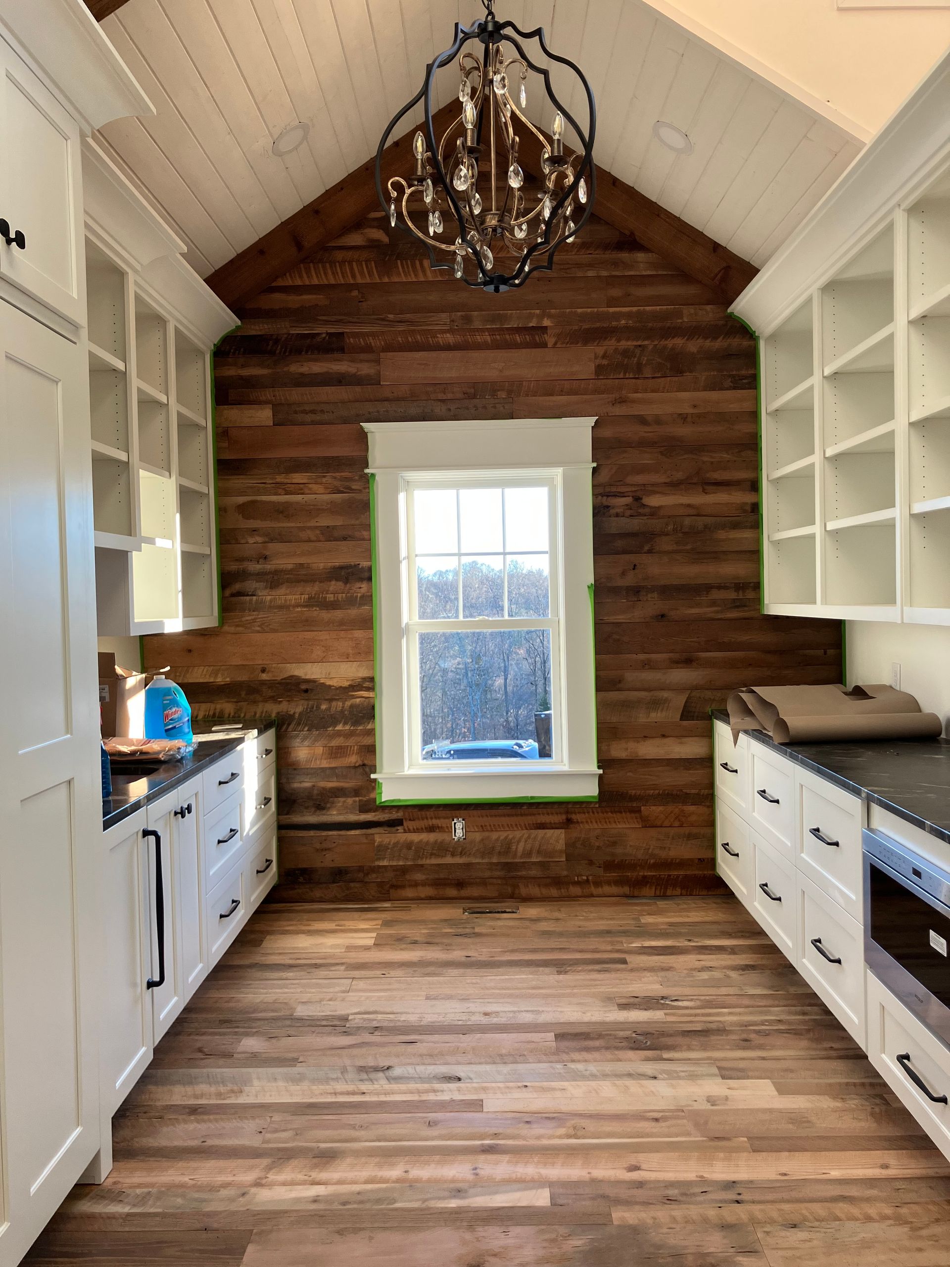 Narrow rustic kitchen with wood walls, white cabinets, black chandelier, and a window at the end.