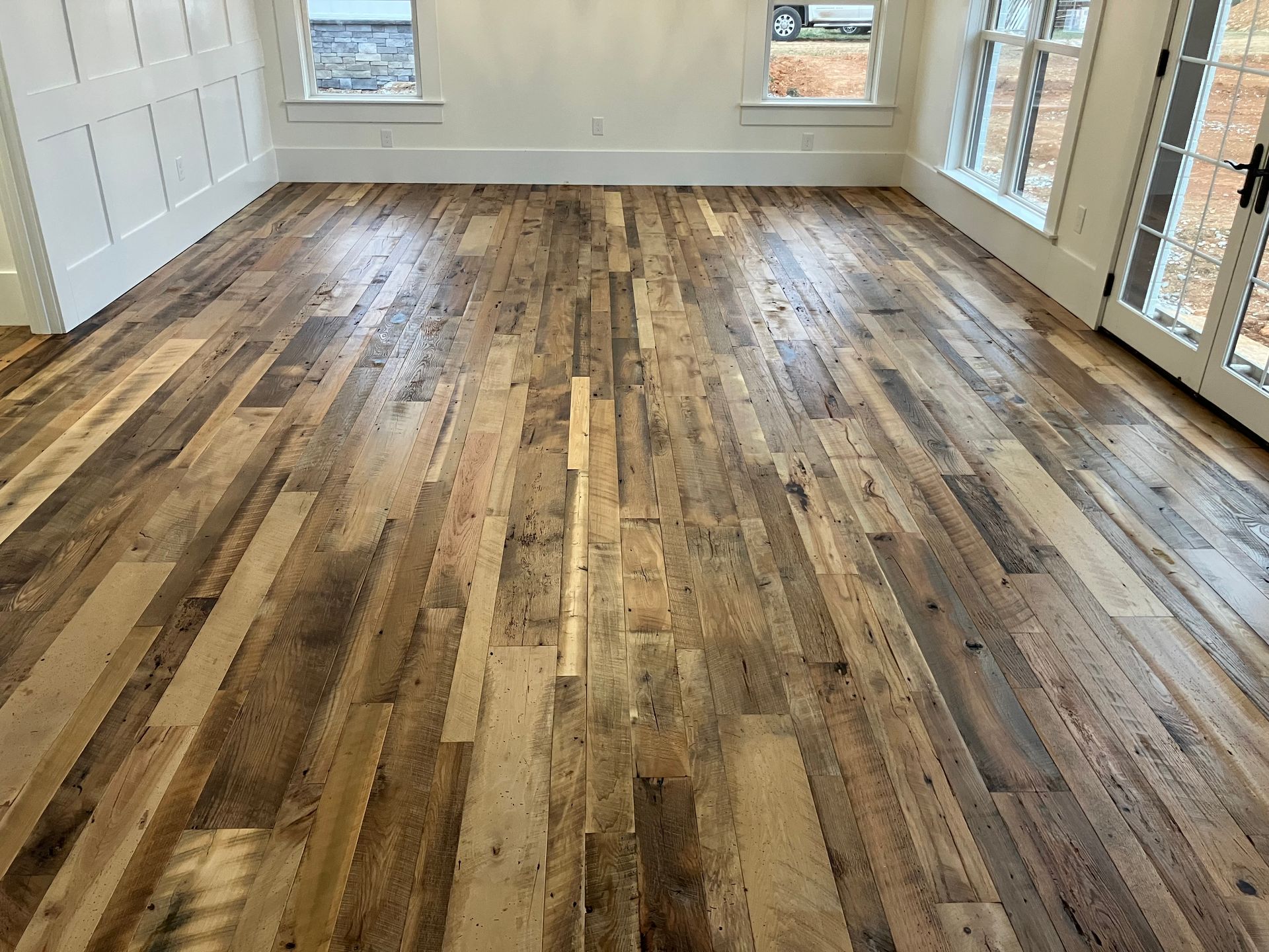Empty sunlit room with worn hardwood floors, white walls, and windows along the back and right sides