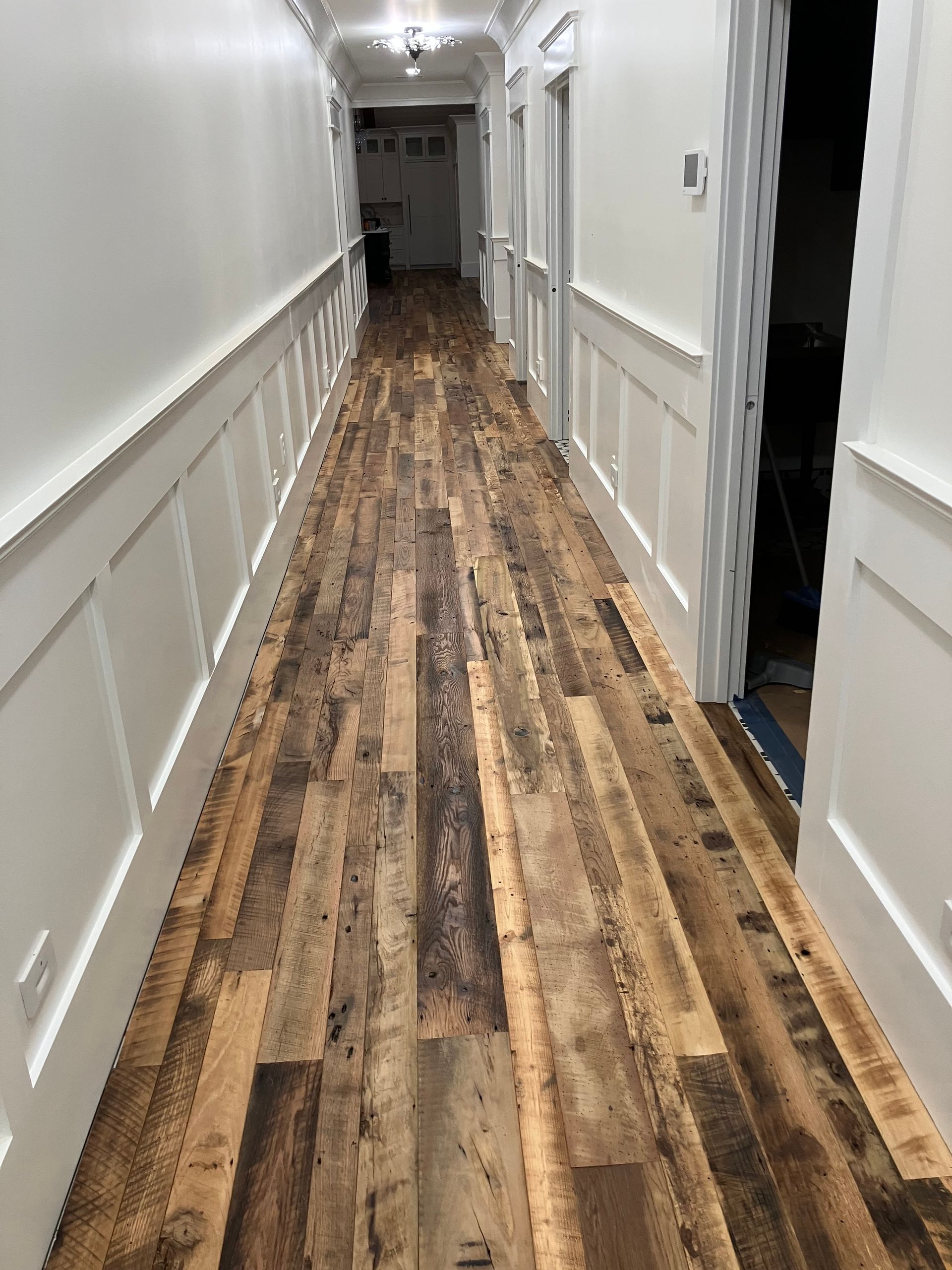 Narrow hallway with white walls, wainscoting, and a worn wood floor leading to a darker room.