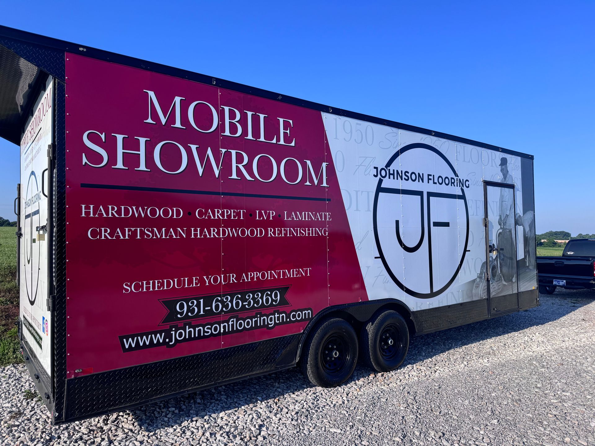 Black mobile showroom trailer with John Ford logo and pink “Mobile Showroom” sign parked on gravel roadside