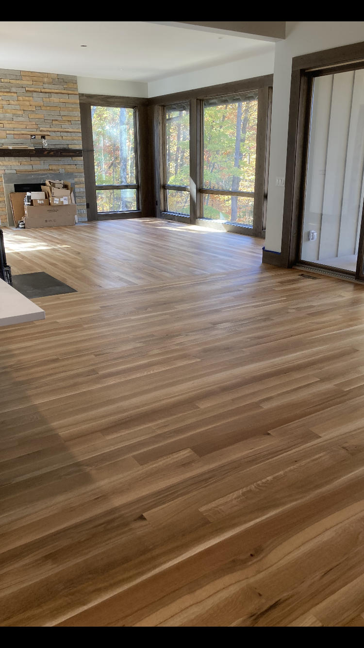 Spacious living room with hardwood floors, stone fireplace, and large windows overlooking trees