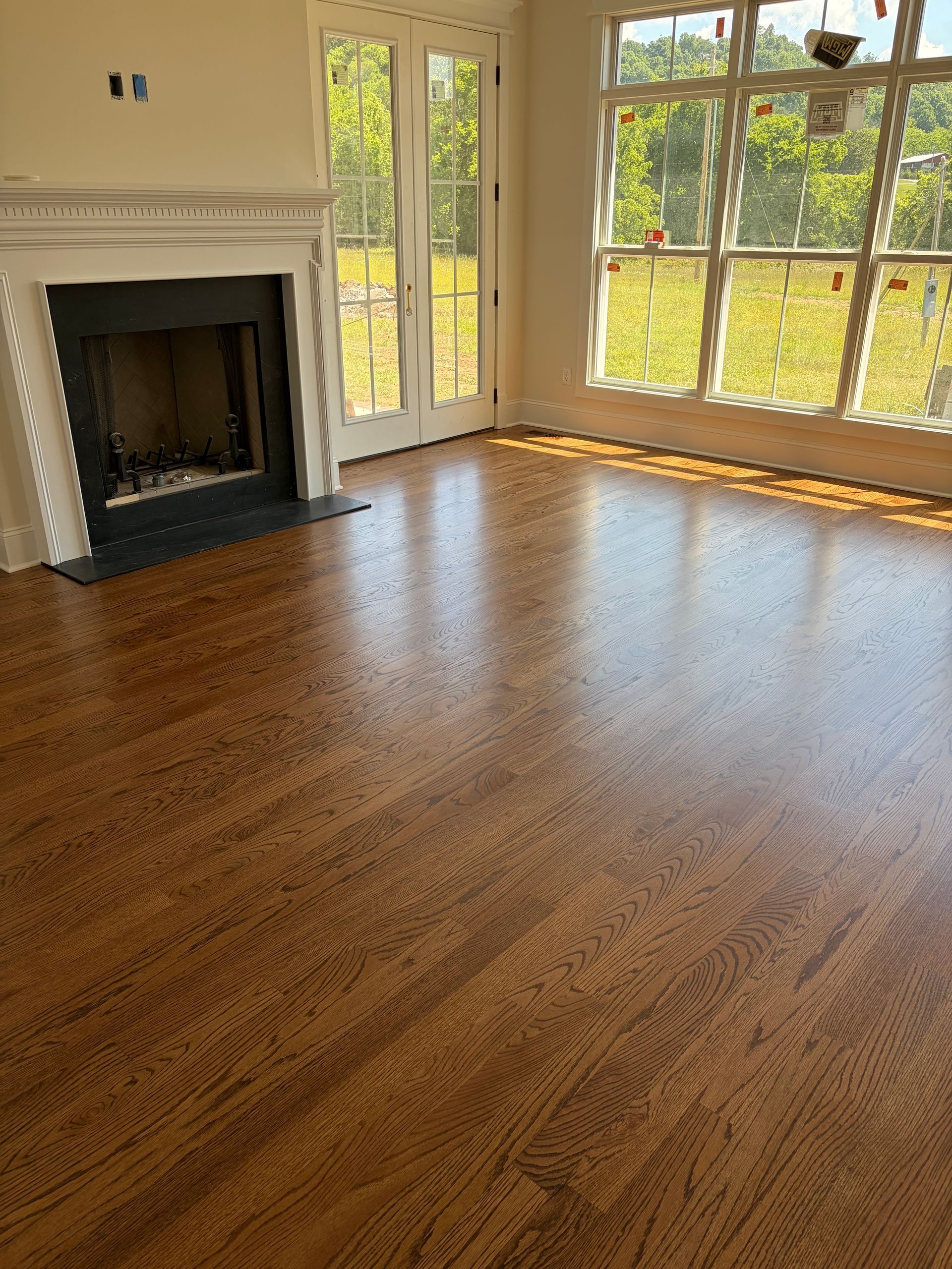 Empty living room with hardwood floor, white fireplace, and large windows overlooking trees.
