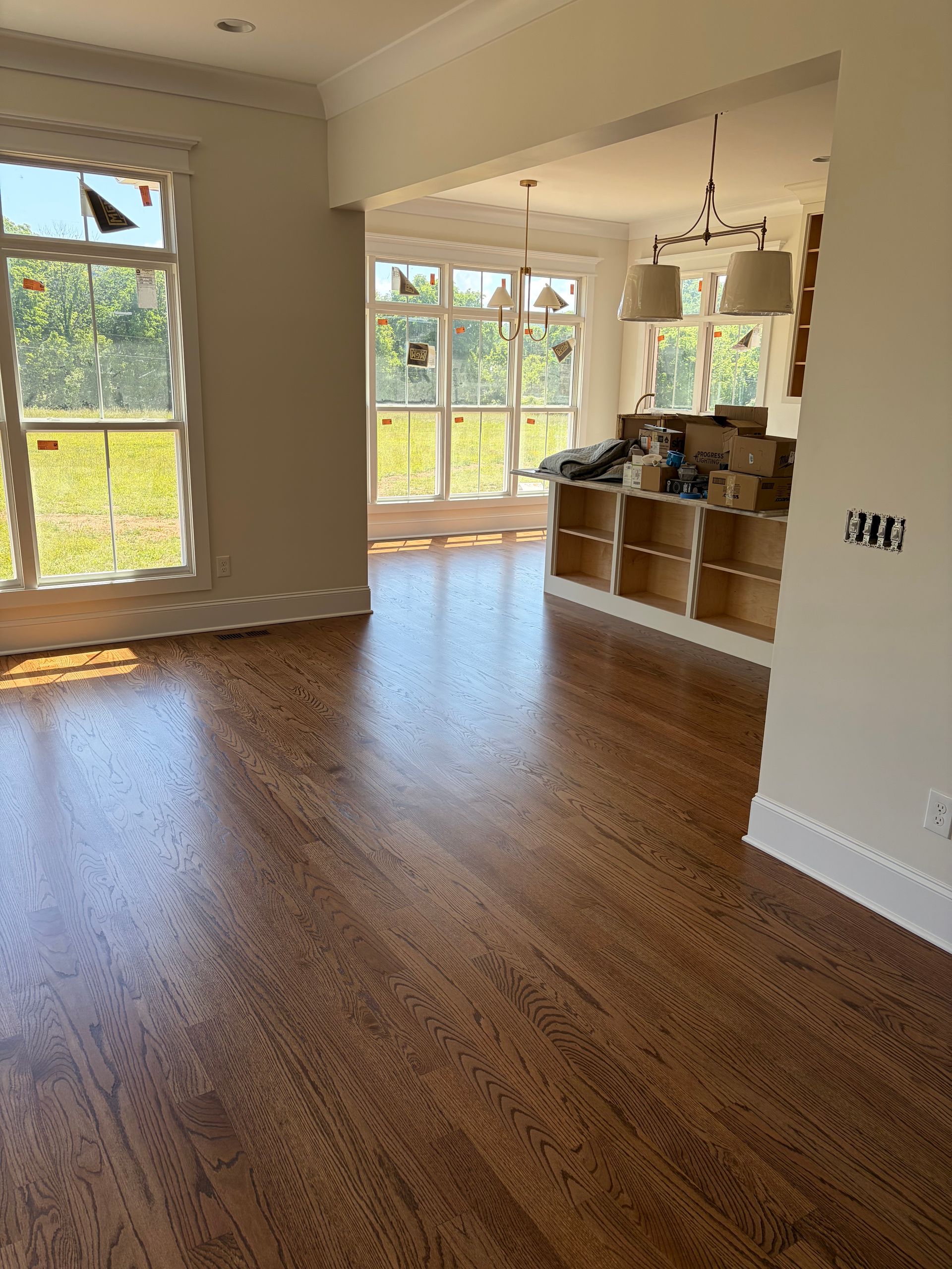 Empty sunlit living room with wood floors, large windows, and a kitchen counter in the background