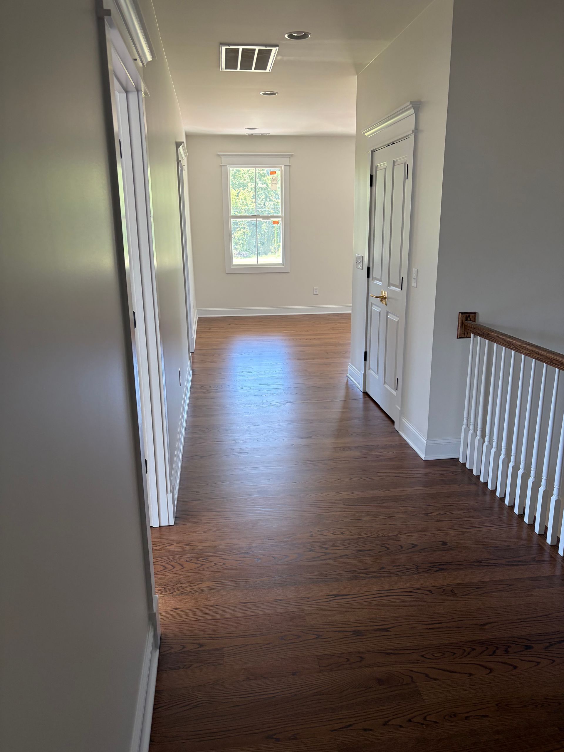 Hallway with hardwood floors, white walls, a window at the end, and stairs with a wooden railing on the right.