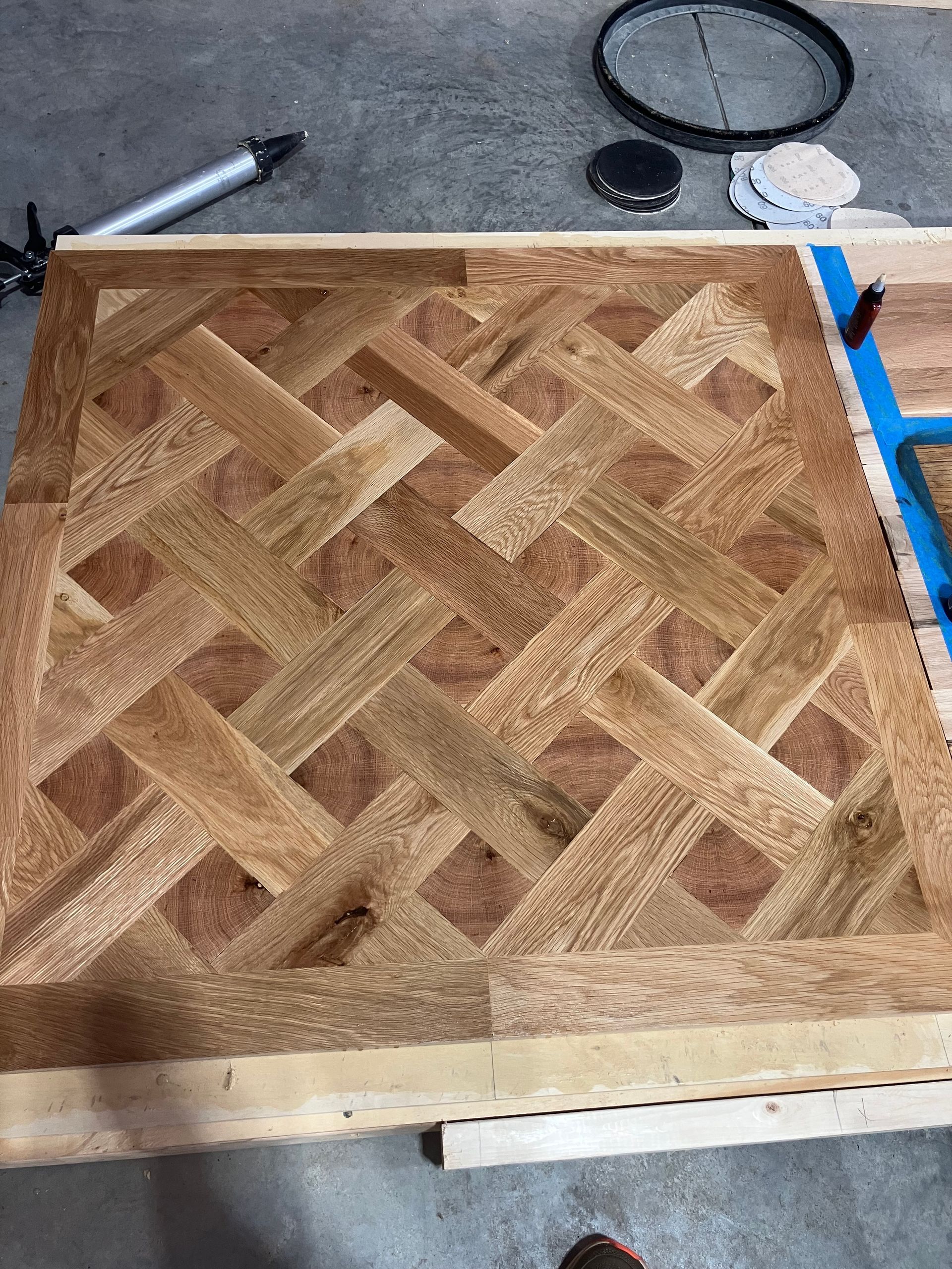 Wooden parquet panel with woven pattern on a workbench, next to tools and a blue clamp.