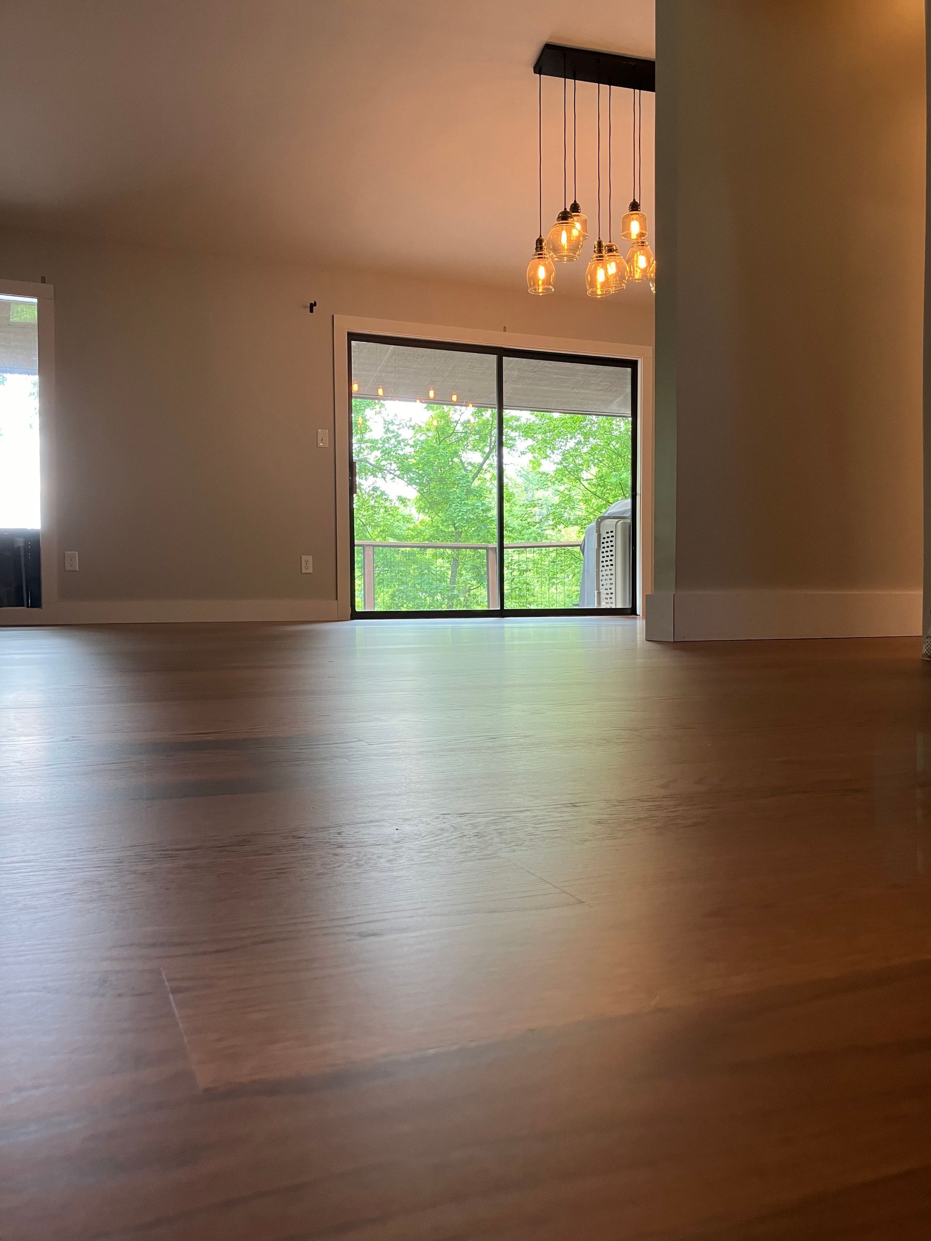 Empty modern room with wood floors, beige walls, hanging lights, and a sliding glass door to green trees