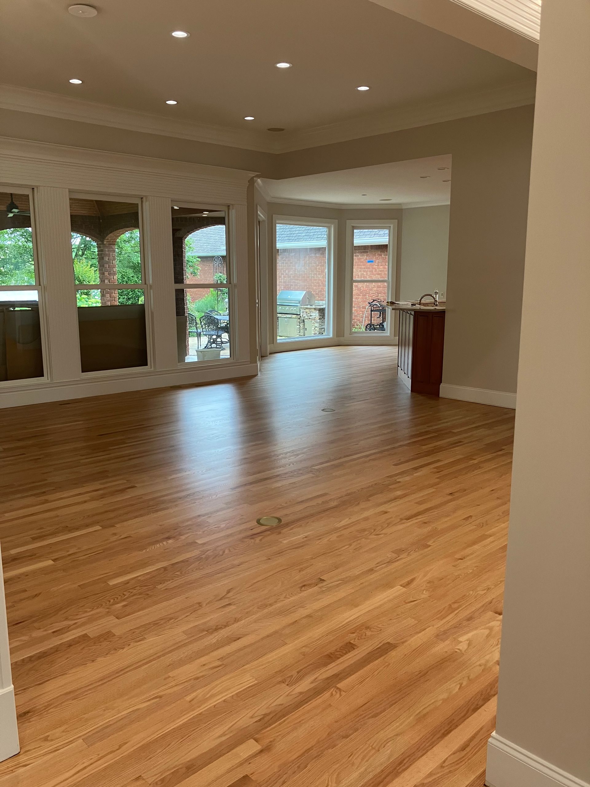 Empty living room with hardwood floors, recessed lights, and tall windows looking onto greenery.